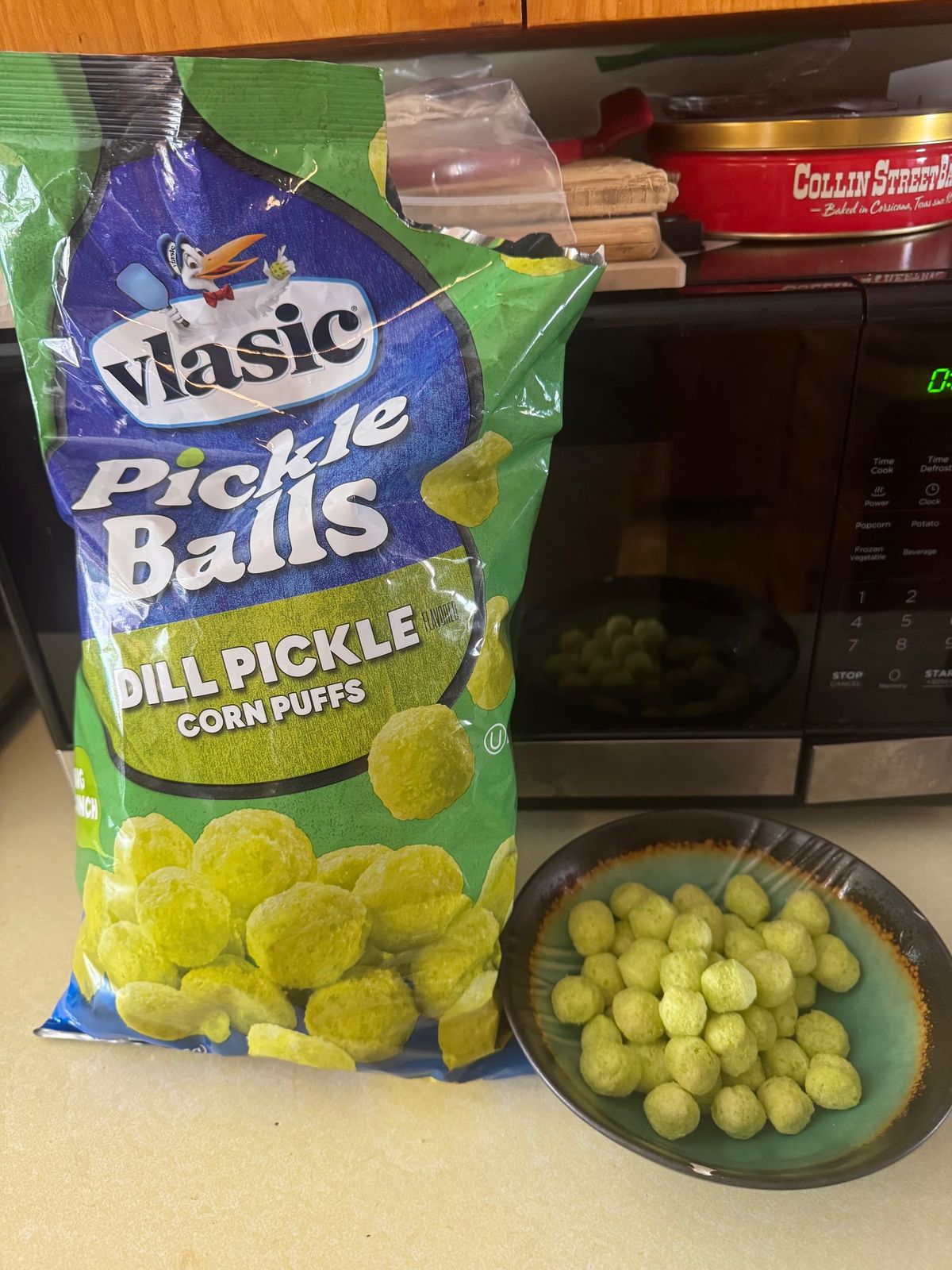 A bag of Vlasic dill pickle corn puffs next to a bowl of ugly green human kibble, in front of a microwave.