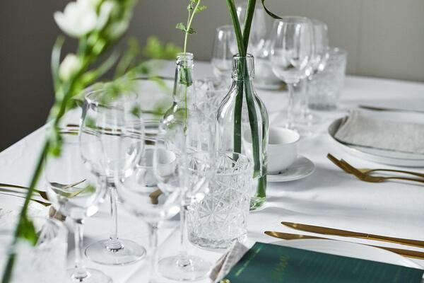 Valoista kattaus: valkoinen pöytäliina, viinilasit, kristalliset juomalasit, lasipulloissa vihreitä oksia ja valkoisia kukkia koristeena, pronssinväriset aterimet ja lautaset joiden päällä kangasservetit. Etualalla kuvassa vihreä menukortti.