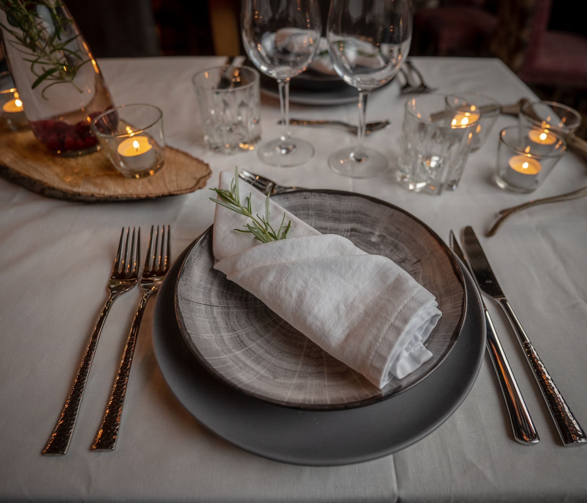 Tunnelmallinen kattaus: kankainen liina, eri sävyiset harmaat lautaset päällekkäin, joiden päällä taiteltu vaalea kangasservetti, jonka sisällä havunoksa. Kahdet koristeelliset aterimet, jälkiruoka-aterimet, viinilasit ja vesilasit. Pöydällä myös tuikkuja läpinäkyvissä lasisissa tuikkukipoissa ja puinen karahka-koristealusta, jonka päällä lsiää tuikkuja ja pullo.