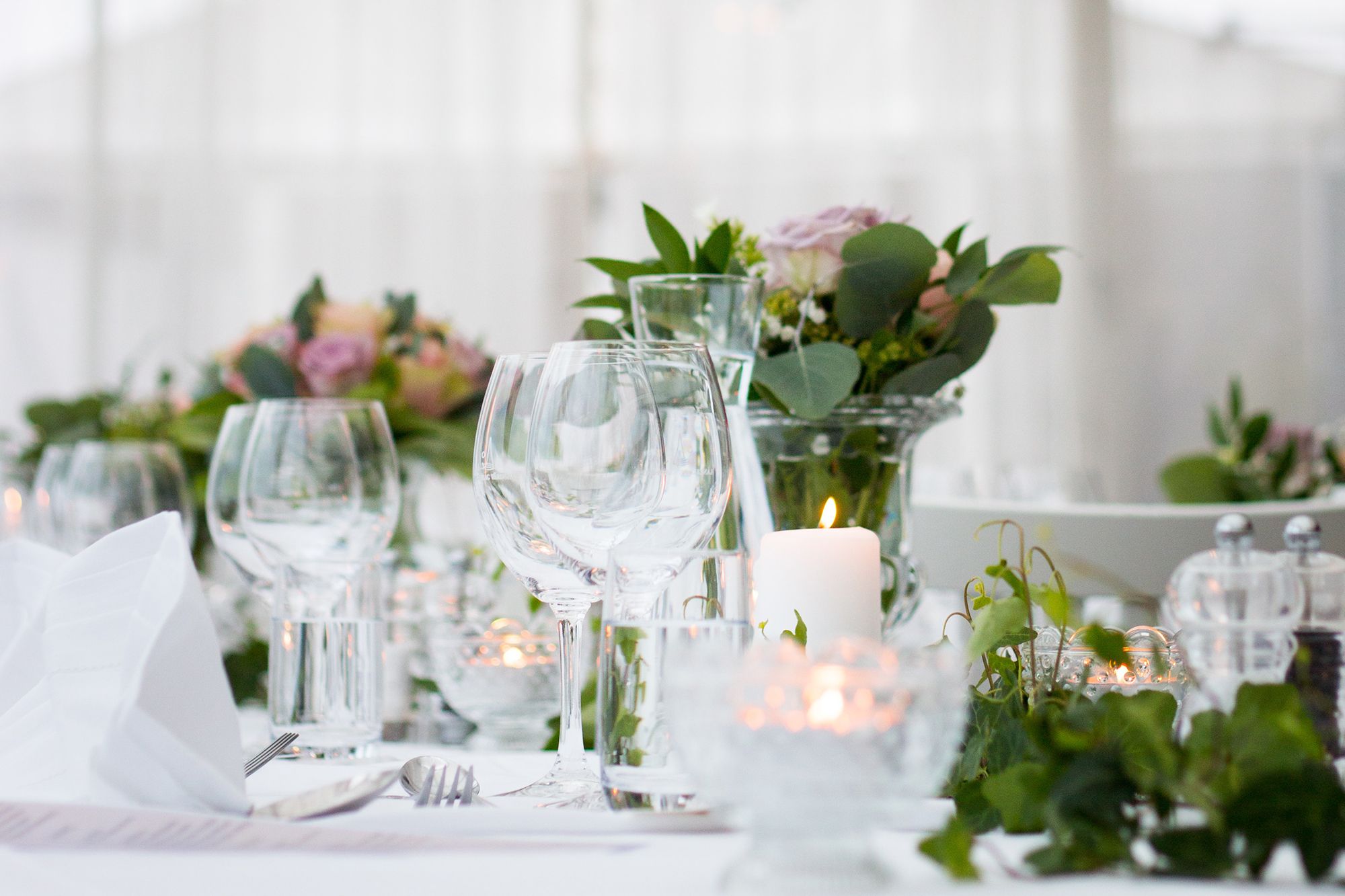 Kaunis ja runsas kattaus: valkoinen pöytäliina, viinilasit, hopeiset aterimet ja valkoiset servetit taiteltuna pystyyn. Lasisissa maljakoissa runsaita vihreitä oksia ja lehtiä sekä roosan värisiä kukkia. Pöydän päällä lisää vihreitä kasveja koristeena. Pöydällä on myös läpinäkyvät suola- ja pippurimyllyt ja muutamia tuikkuja palamassa lasikipoissa ja pöydällä palaa yksi valkoinen pöytäkynttilä.