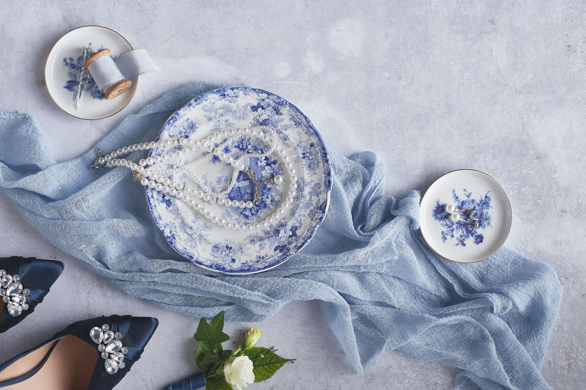 Kuvituskuva: tummansiniset hääkengät, ruusu sekä kolme sinertävää lautasta vaaleansinisen liinan päällä, joissa helmikoruja häihin