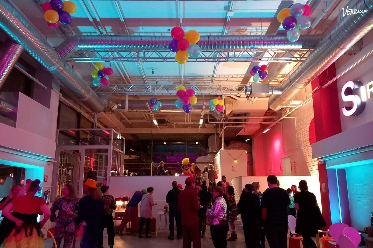 Korkea juhlatila, jossa ihmisiä, tunnelmallinen värivalaistus sekä ilmapallokoristeita