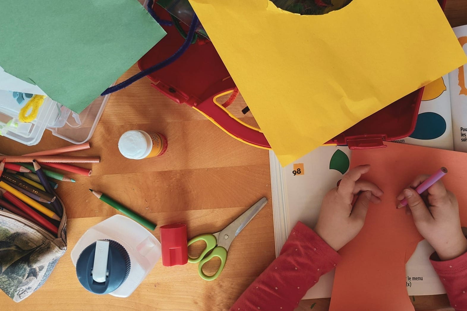 A child drawing on orange paper at a wooden table with craft supplies