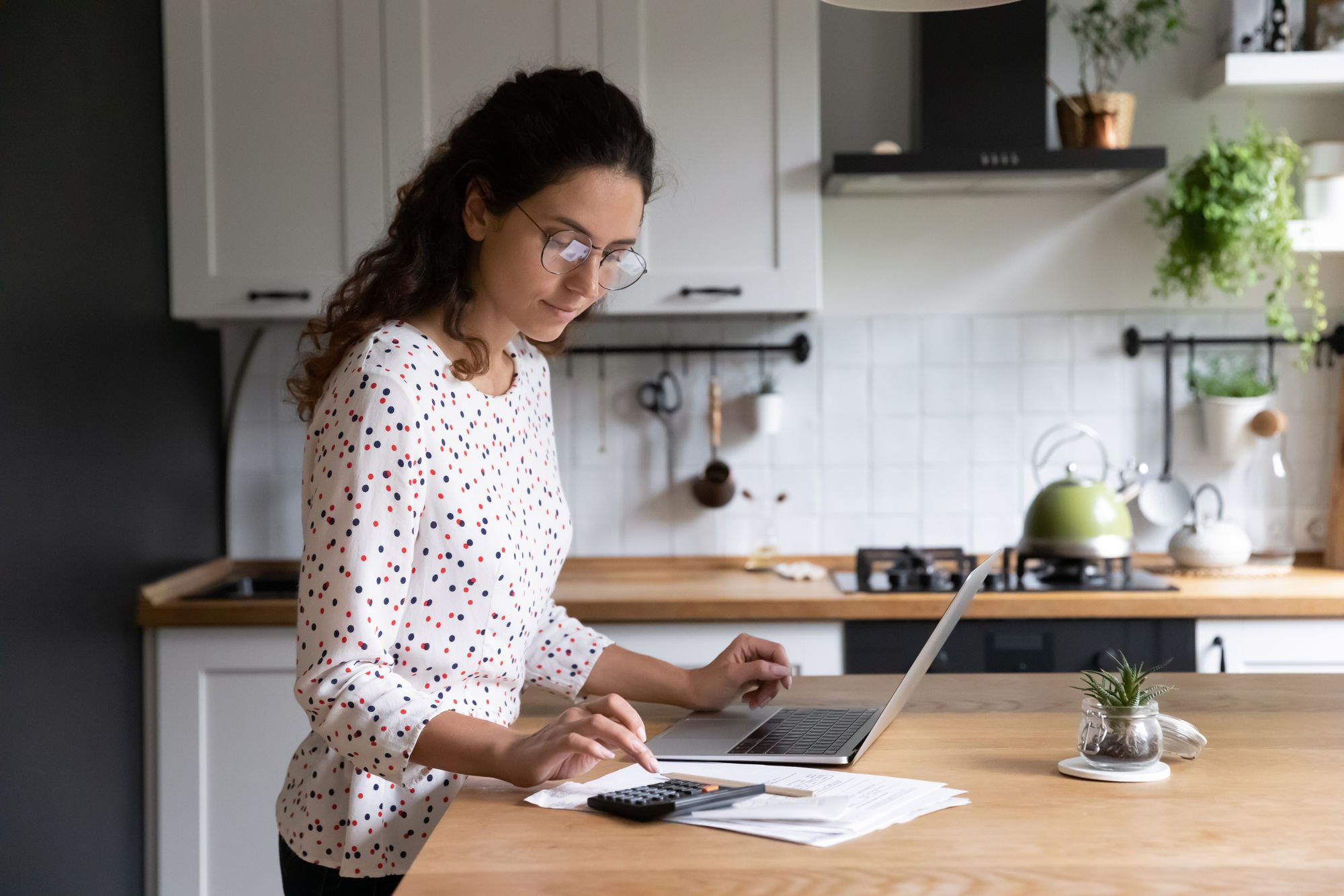 A woman stands at her kitchen counter using a laptop and calculator to determine if she can afford the mortgage origination points for her home purchase.