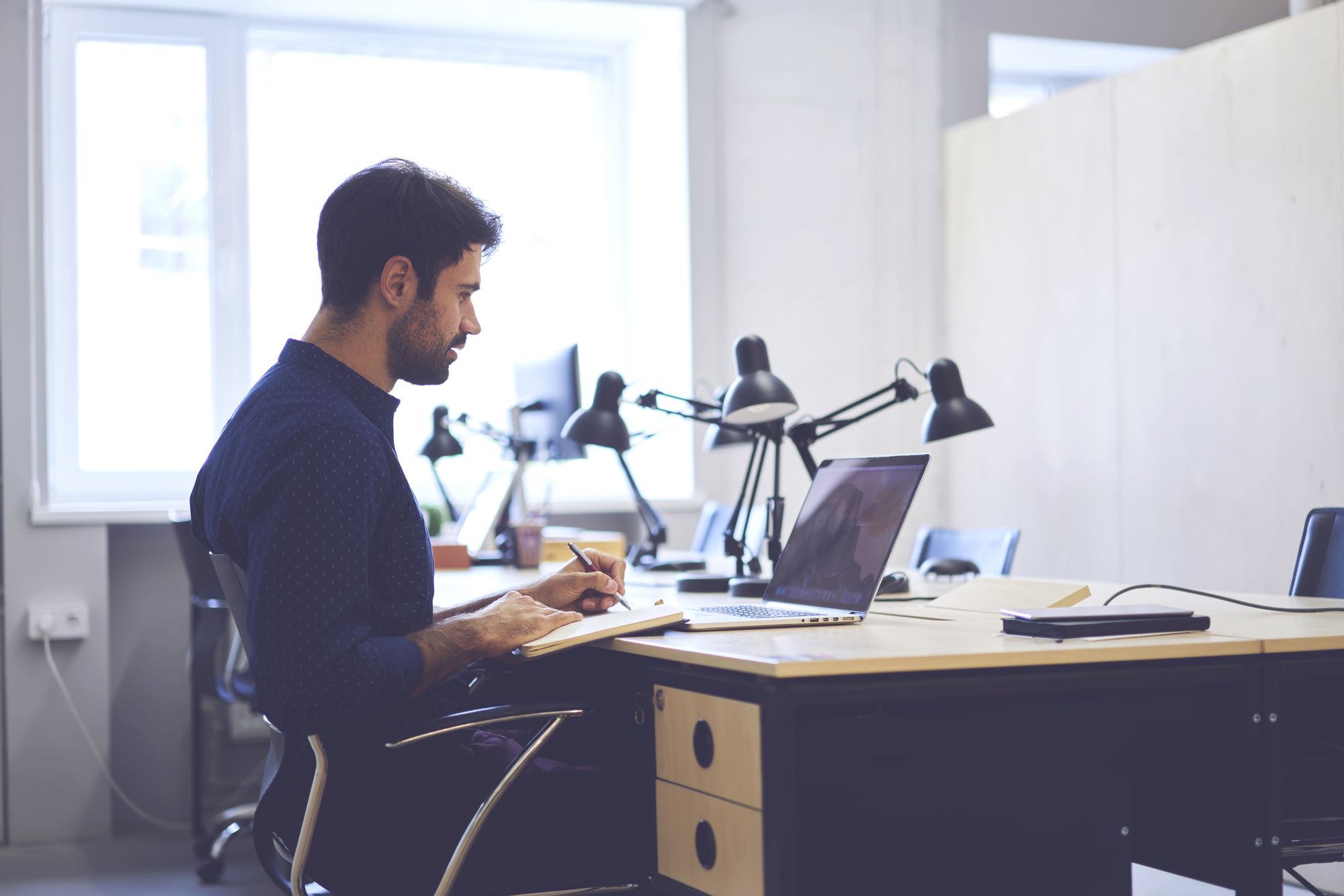 A male real estate agent sits at their desk looking at their laptop while taking notes in a journal.
