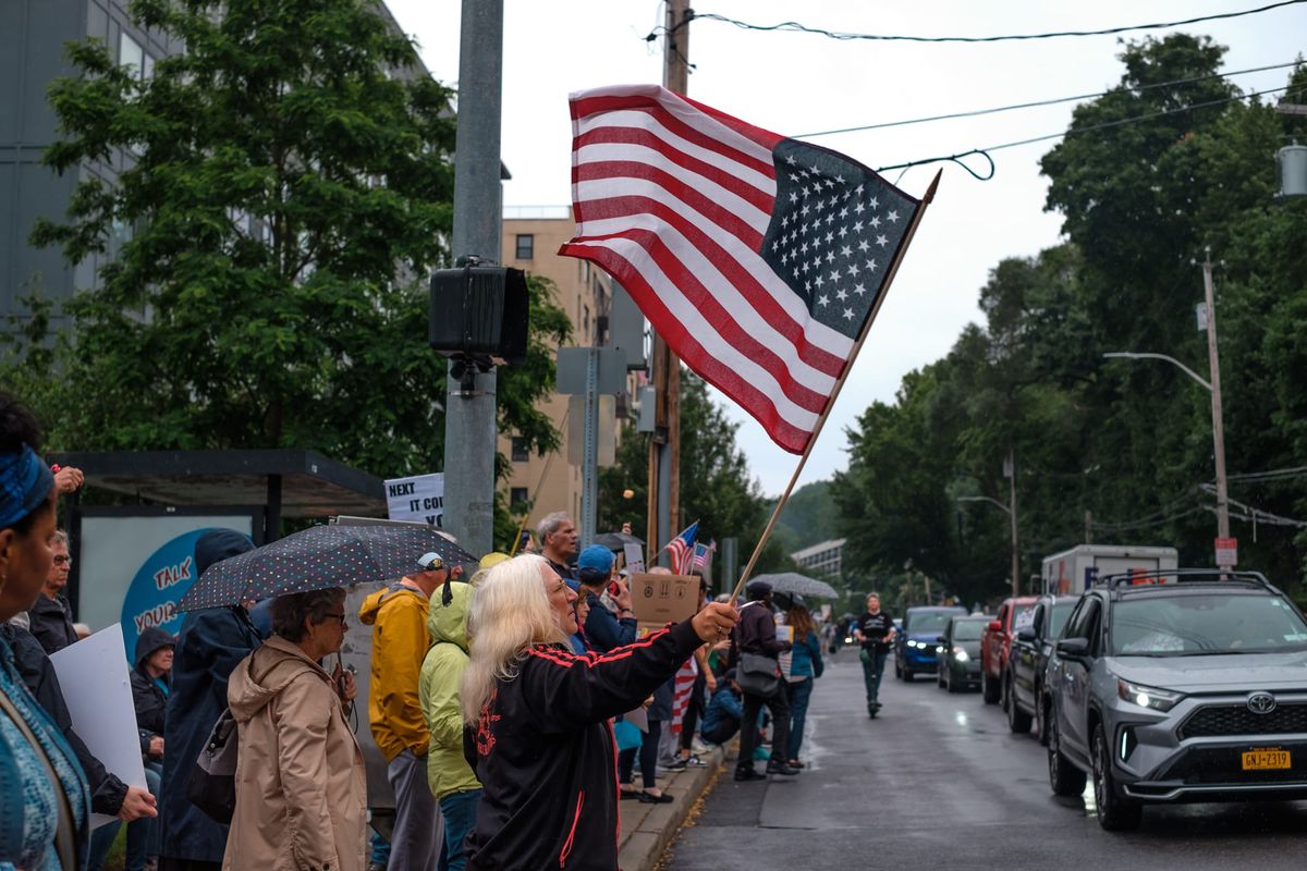 A crowd gathers along the side walk as a woman stands at the edge waving a giant American flag. The sky is cloudy and rainy. A line of cars stretch down the street where more people can be se