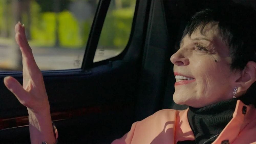 An ageing Liza Minnelli in an orange jacket and black turtleneck looks out the window from inside a car.
