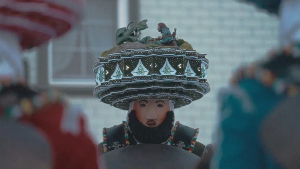 A person wears a costume with a bearded mask and elaborate headdress made of carved wooden scultures.