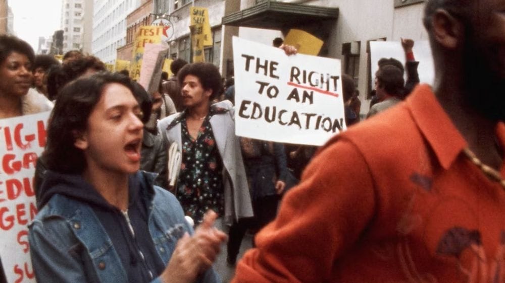 A woman stands amid a protest where a man holds a sign reading The Right to an Education.