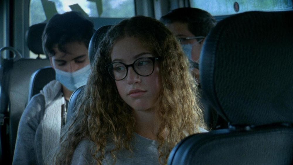 A teenage girl with long curly hair sits on a school bus as other students behind her wear facemasks.