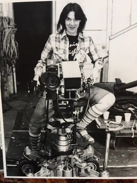 A young woman with shaggy dark hair wearing plaid and denim shorts with striped socks sits behind a movie camera.