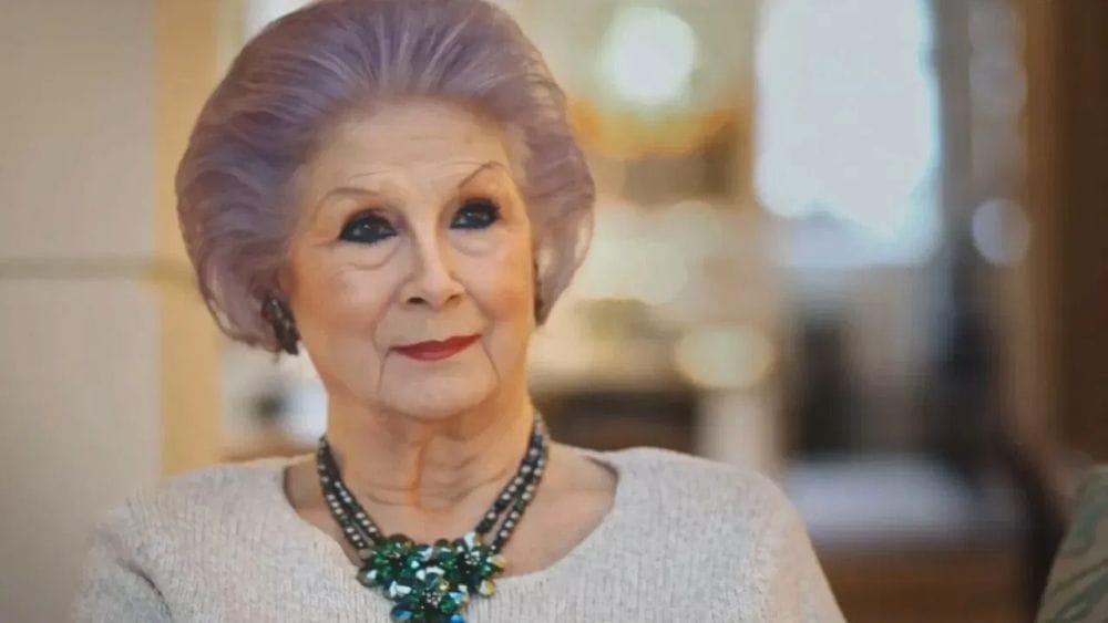 An older woman with dignified but purple hair, deep eye-shadow and a large necklace.