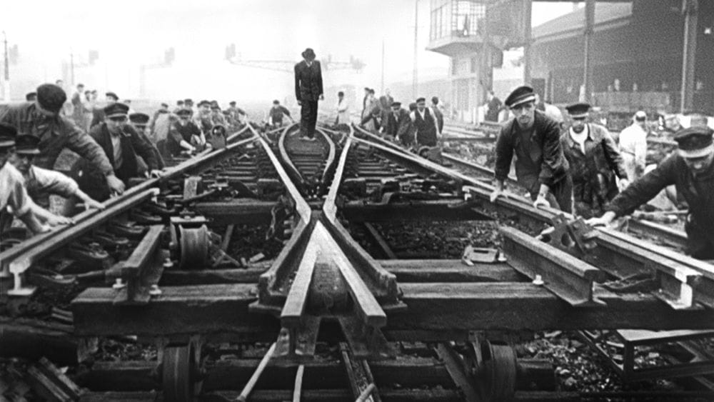 A man in a suit stands in the middle of train tracks and workers flank him on either side putting them into place.