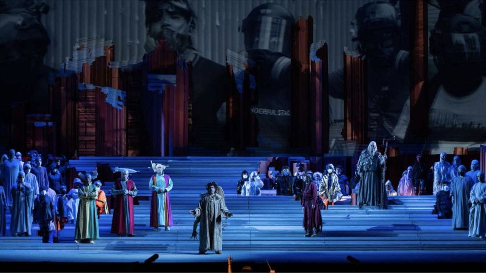 An opera is performed on stage by actors on stairs in colourful costumes against projections of modern day protests.