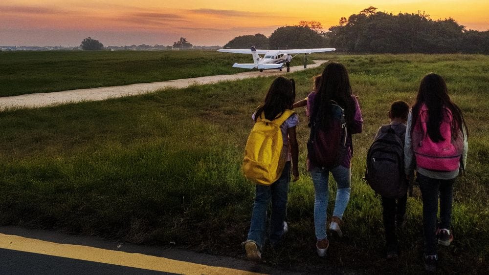 Four siblings with backpacks walk towards a small airplane.