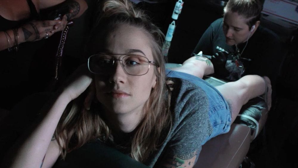 A young woman in glasses gets a tattoo on her leg.