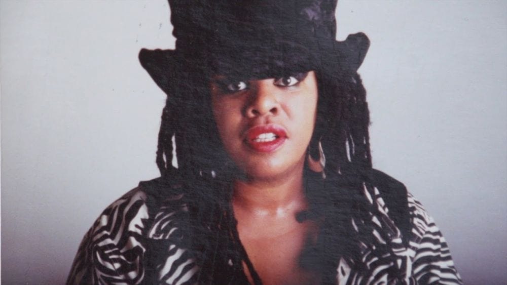Q Lazzarus, a black woman with long dreaded hair, red lipstick, a zebra-print blouse and tophat stares at the camera.