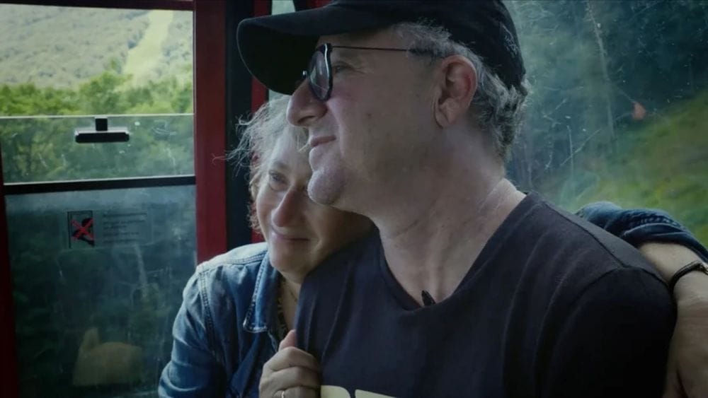 Heidi sits next to Charlie with her arm around the back of his shoulders.