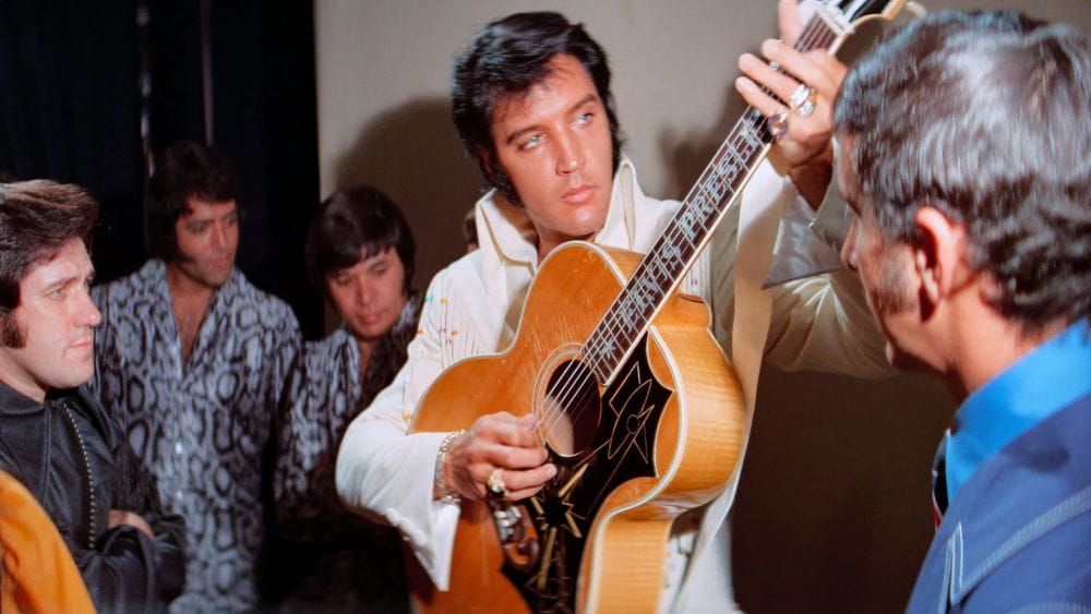 Elvis Presley in a white jumpsuit tunes a guitar backstage surrounded by backup singers and musicians.