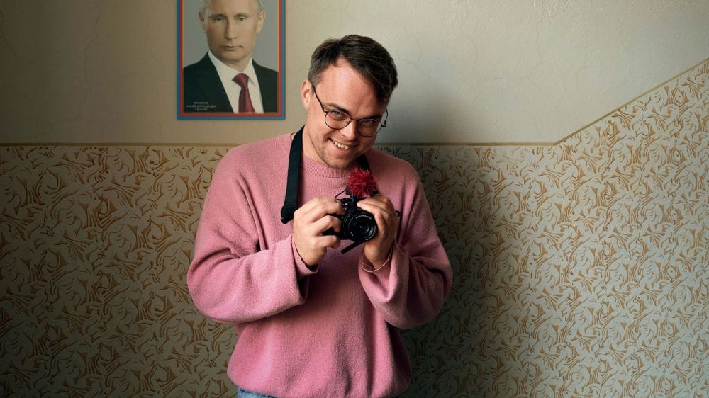 A man in glasses and a pink jumper holds a camera, standing in front of a photo of Vladimir Putin.