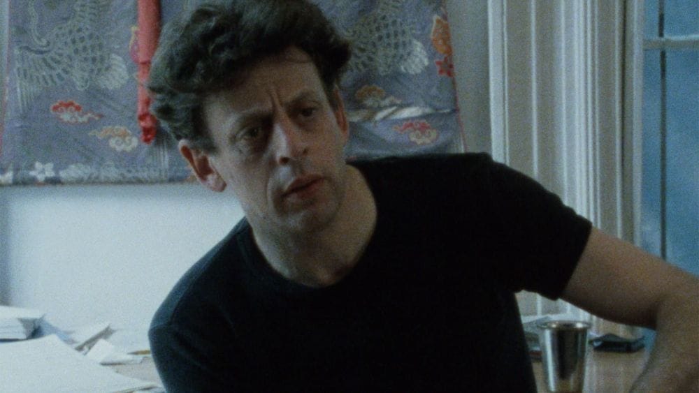 Robert Wilson in a black shirt looking frustrated in an office with art on the wall and a desk covered in papers.