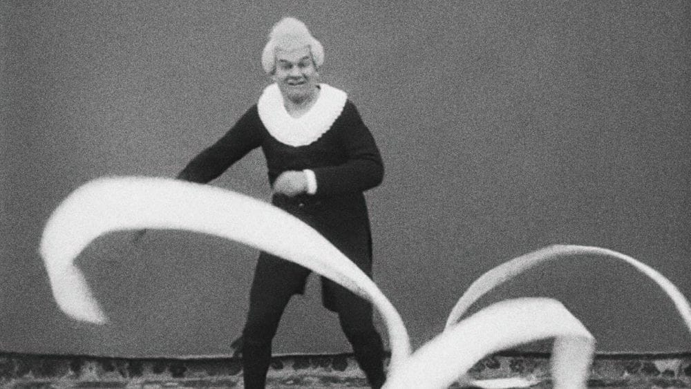 A man in black with a white collar and wig waves a large white ribbon as he dances.
