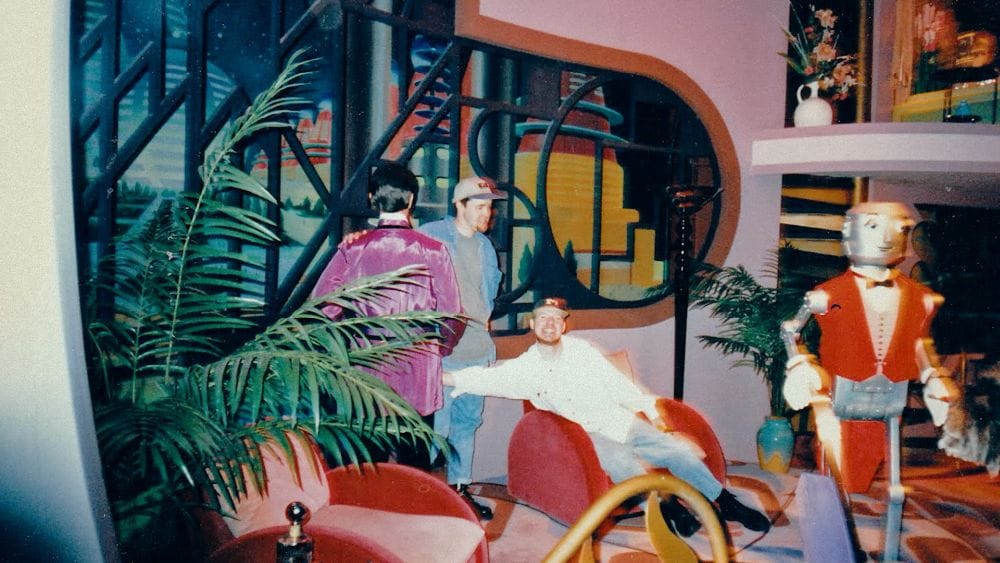 Two men from the '90s, lounging in a room of animatronics in a room painted pink and 1950s furniture.