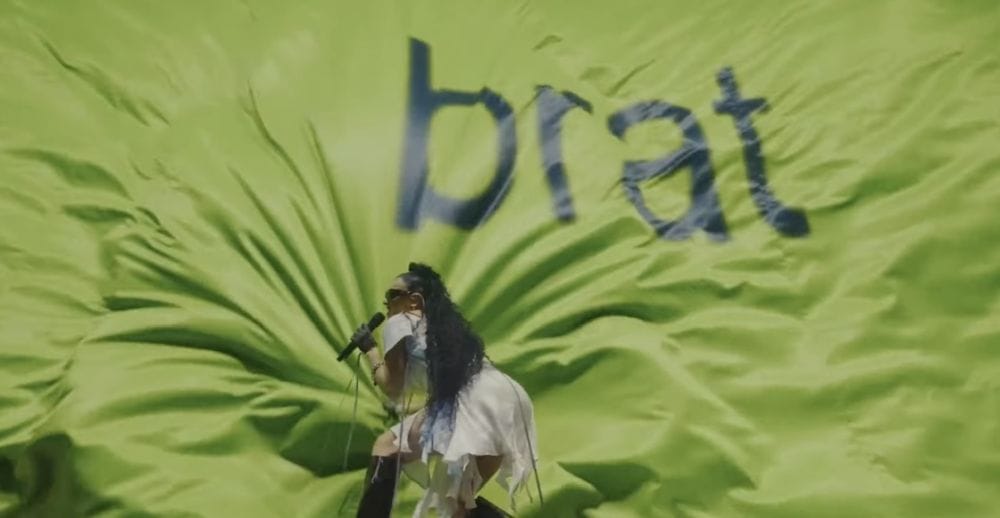 Charli XCX in a white dress and hip-length curly hair performs in front of a green curtain that reads the word brat.