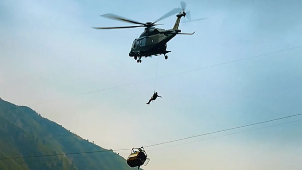 A boy hangs from a wire out of a helicopter above a tipped cable car.