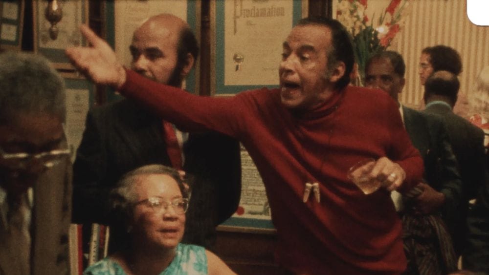 Richard Bruce Nugent wearing a red turtleneck gesticulates at a party guest.