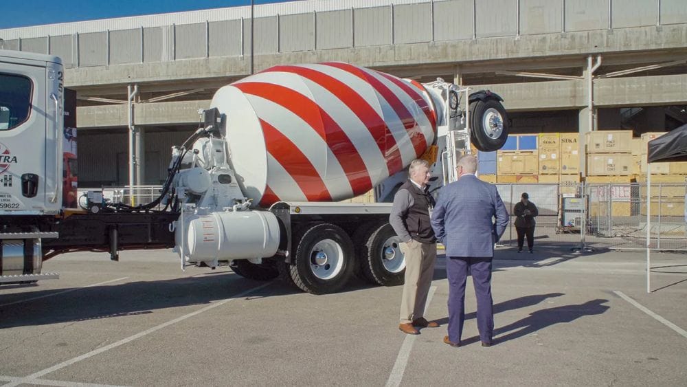 Two men in business clothes stand next to a red-and-white striped cement mixer.