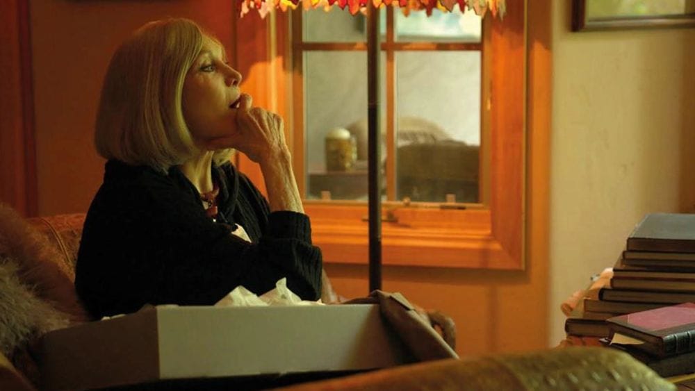 An Ageing Kim Novak sits in a room surrounded by boxes and books as she looks off into the distance.