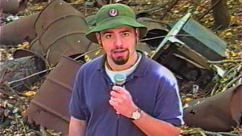 A teenage reporter with a goatee stands in a dump reporting the news.
