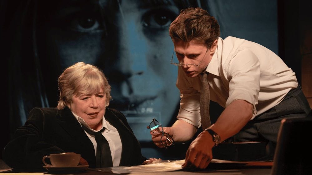 Marianne Faithfull in her 70s sits in front of a large projection of her younger self while a young man in white shirt, tie and glasses shows her documents.