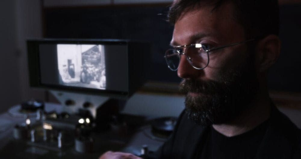 A man with beard and glasses sits in front of a editing machine that is playing a black and white movie.