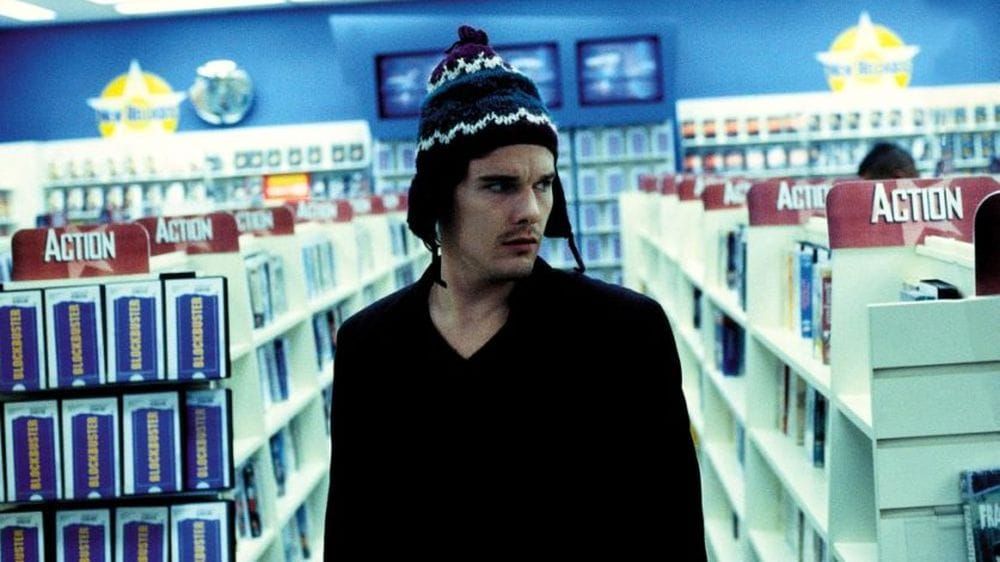 Ethan Hawke in a beanie and black shirt walks down the action aisle of a Blockbuster video.
