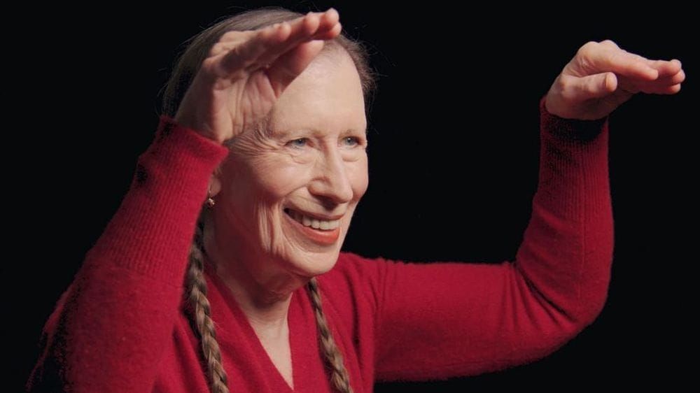 Meredith Monk stands in a red sweather and her hair in pigtails, smiling while elevating her hands.