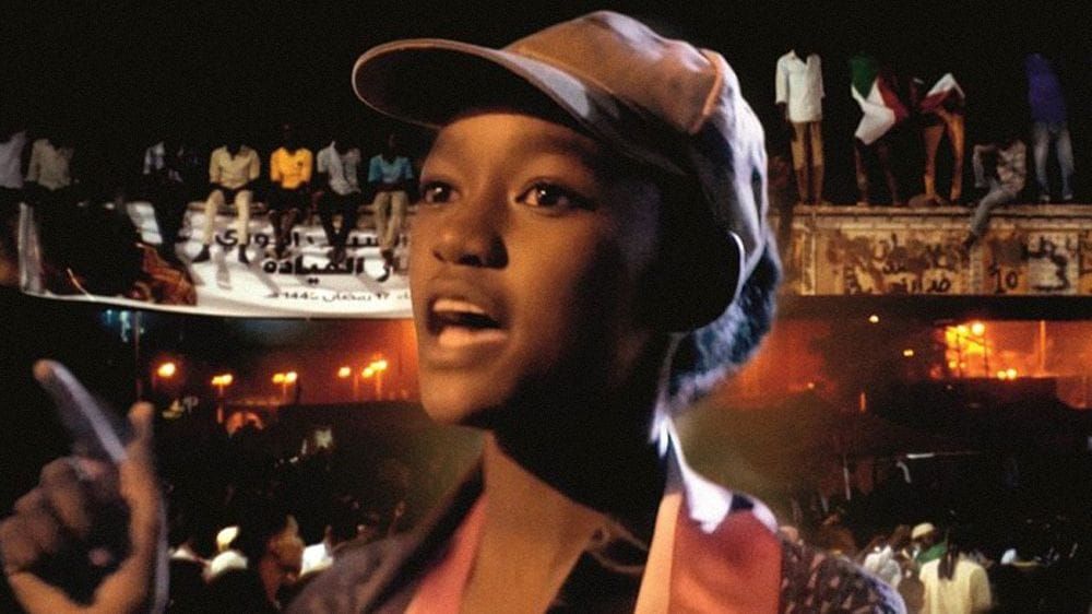 A yound Sudanese woman in baseball cap stands in front of a group of men, rapping about freedom.
