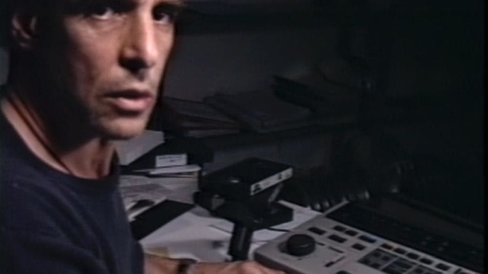 A man looks at the camera, turning from his desk covered in technical equipment.