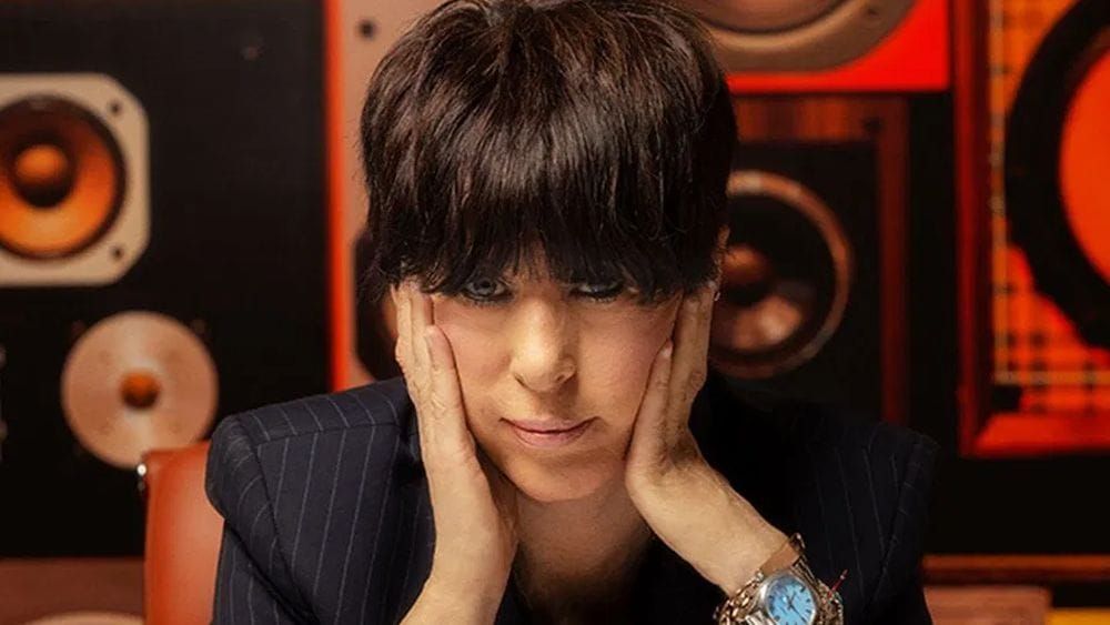 Diane Warren sits in front of a wall of speakers and stares directly into the camera with her hands on her face.
