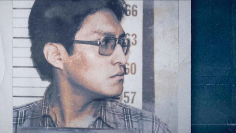 An Apache man in glasses and a plaid shirt stands in front of a police height chart looking to his side.