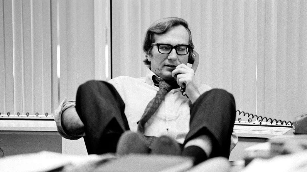 Black and white photo of a man in white business shirt, tie and dark slacks, leaning back in a chair while on the telephone.