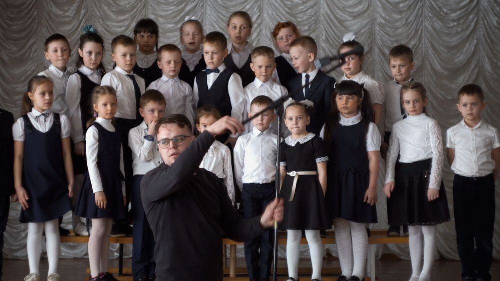 A teacher in black adjusts a microphone in front of a classroom choir of young children.