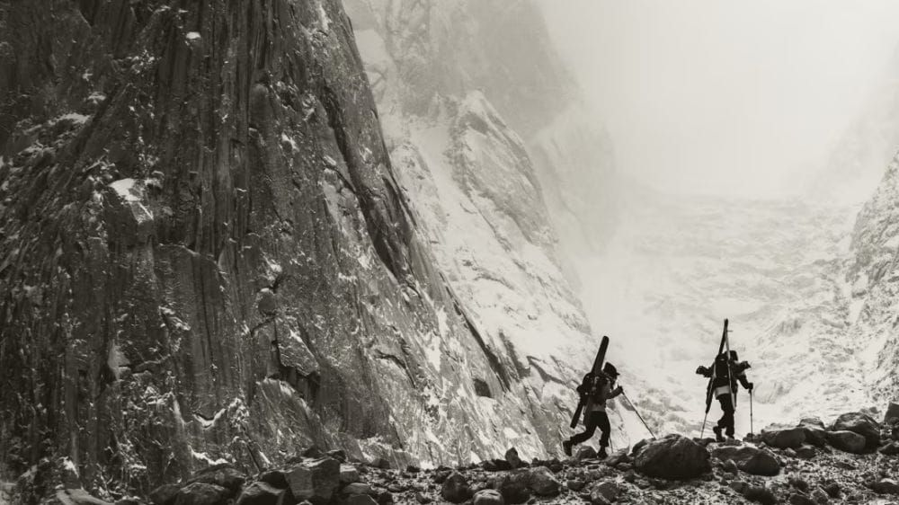 Two people hike with skis on their face as massive mountain faces appear behind them.