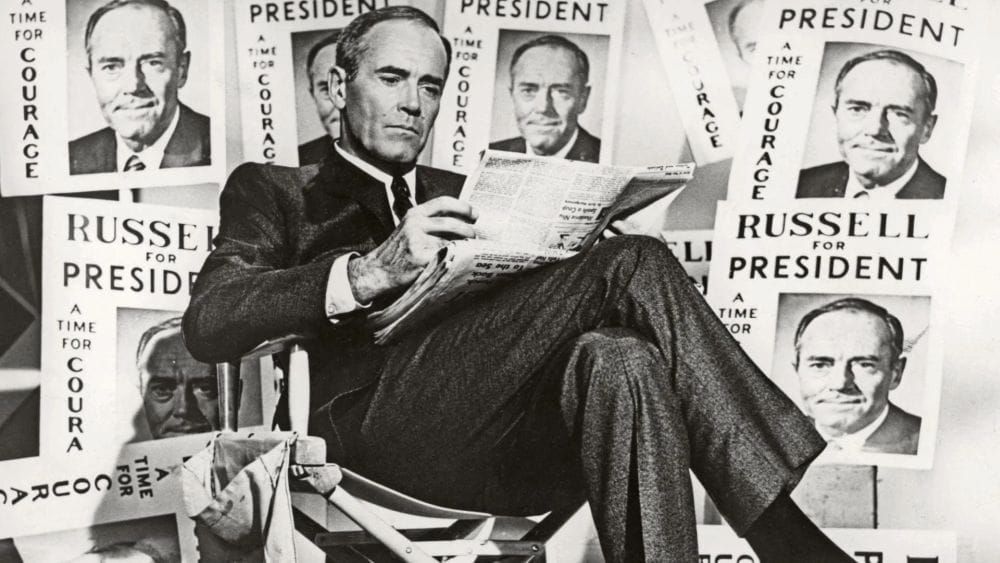 Henry Fonda in a suit and tie sits cross-legged reading a newspaper surrounded by signs that read Russell for President, a time for courage.