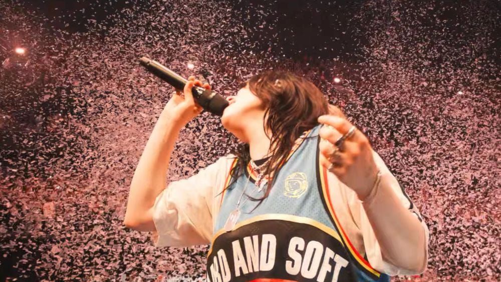 Billie Eilish performs on stage wearing a basketball jersey that reads "Hard and Soft" while confetti falls around her.