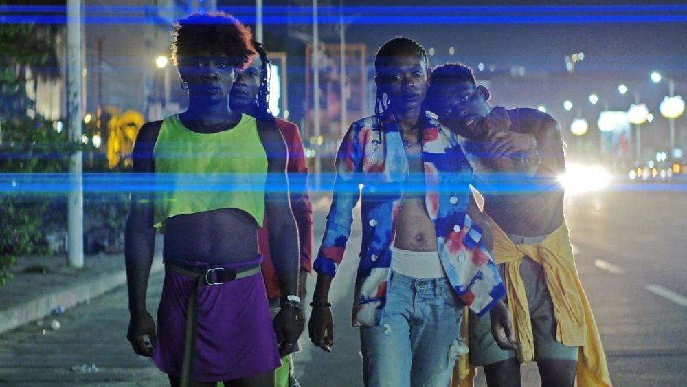 Four black dancers wearing brightly-coloured clothes on a street as car headlights shine around them.