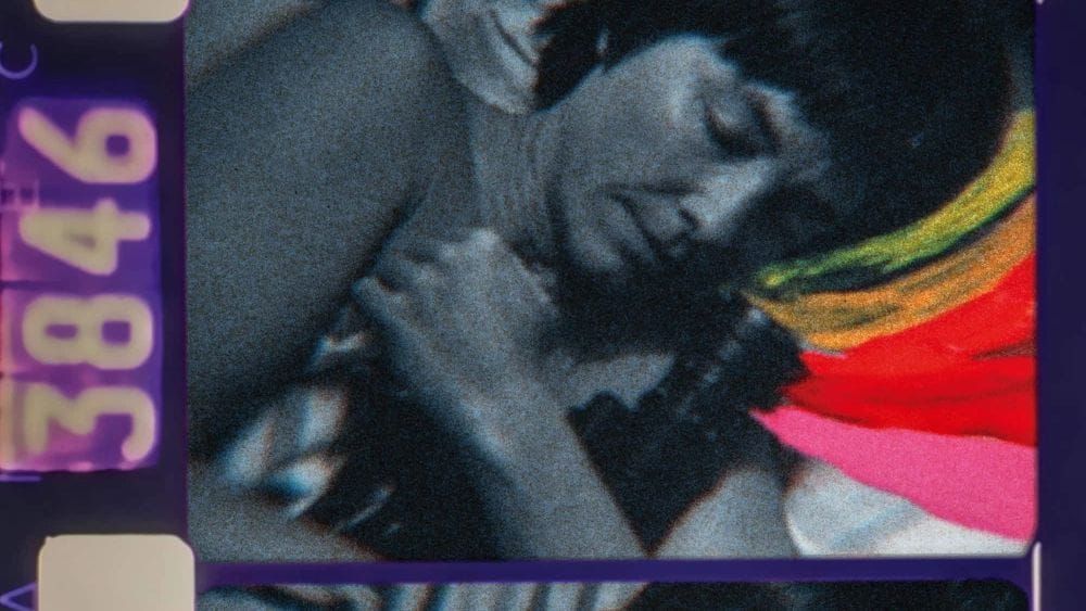 A frame of black and white celluloid showing woman holding an old bolex camera as yellow, orange, red and pink streaks emerge from its lens.