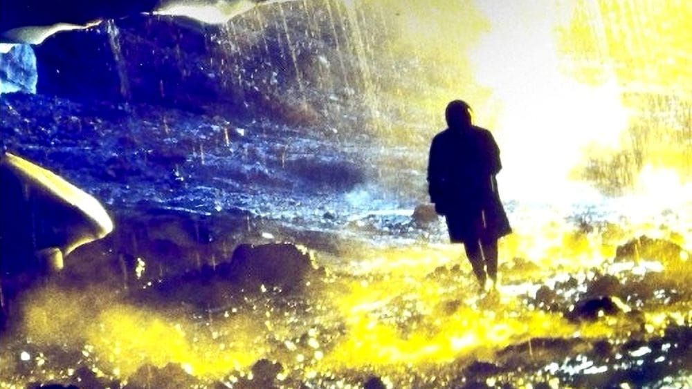 A silhouetted figure walks through a cave dripping with water coloured by yellows and blues.