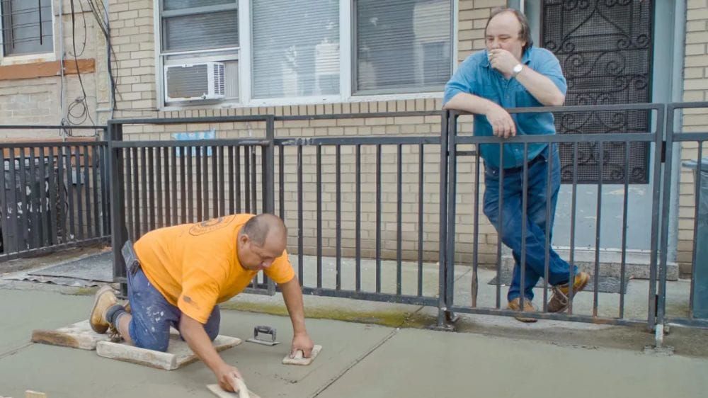 A tradesman smoothes wet concrete on a sidewalk while a man watches while smoking a cigarette.